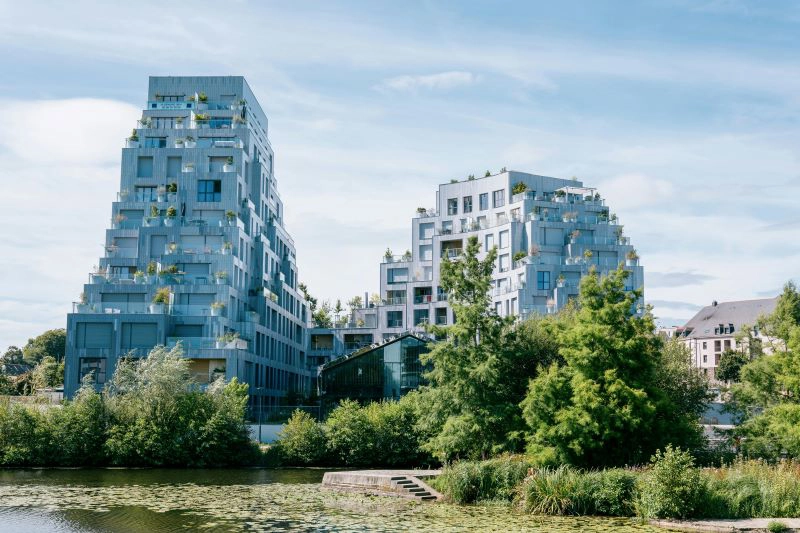 Sous un un ciel bleu clair, deux bâtiments résidentiels situés sur les bords de la Vilaine à Rennes, dans le quartier de Baud-Chardonnet. Il s'agit d’un ensemble d’immeubles modernes et emblématiques de l’architecture contemporaine rennaise, souvent surnommés “les pyramides végétalisées” en raison de leur forme en gradins et de la végétation intégrée aux terrasses.